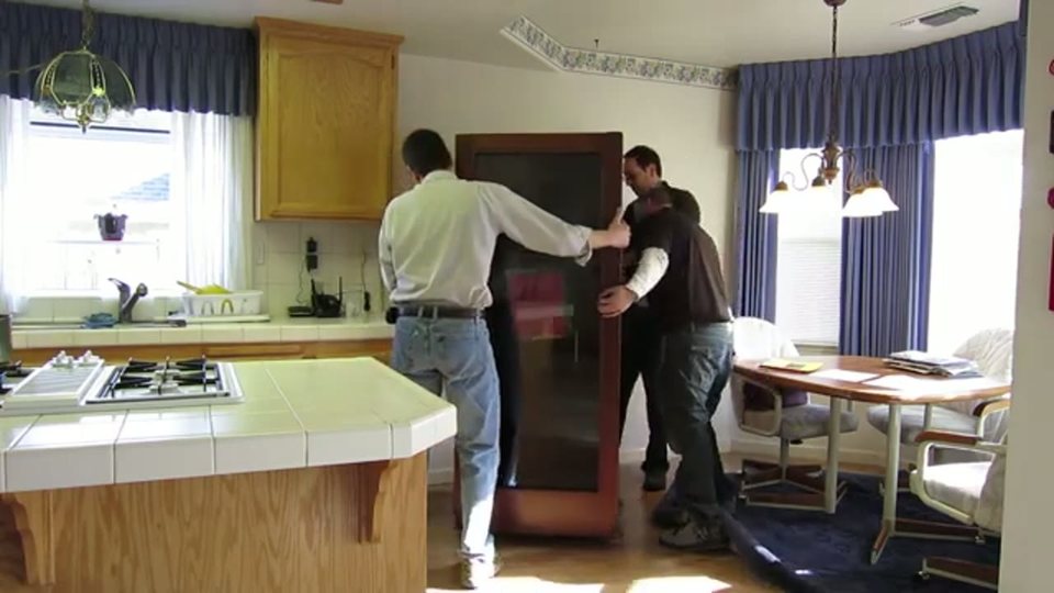 three movers guiding a tall shrink-wrapped wardrobe through a kitchen